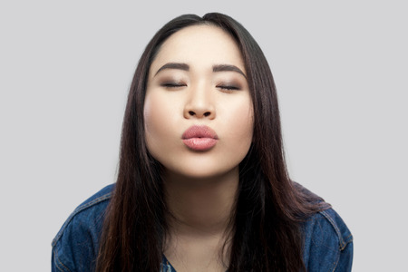 Portrait of in love beautiful brunette asian young woman in casual blue denim jacket with makeup standing with closed eyes and sending kiss. indoor studio shot, isolated on light grey background.の写真素材