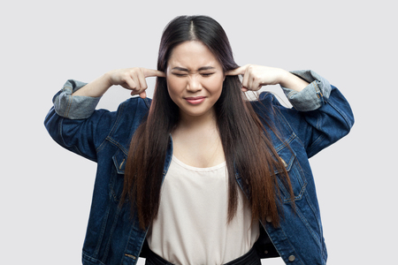 Portrait of nervous beautiful brunette asian young woman in casual blue jacket with makeup standing and putting finger on ears and don't want to hear. studio shot, isolated on light grey background.の写真素材