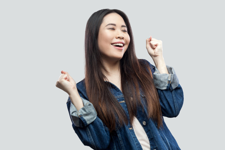 Portrait of happy surprised winner beautiful brunette asian young woman in casual blue denim jacket with makeup standing and celebrating her victory. studio shot, isolated on light grey background.の写真素材