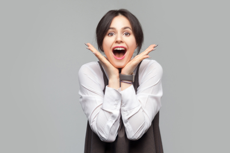 Excited beautiful young woman in white shirt and brown apron with makeup, collected hair standing with hands touching face and looking at camera with surprised face. studio shot on grey background.の写真素材