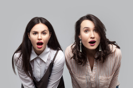 Unbelievable news. Closeup portrait of shocked beautiful brunette girls in casual style standing together and looking at camera with open mouths and big eyes. studio shot, isolated on grey backgroundの写真素材