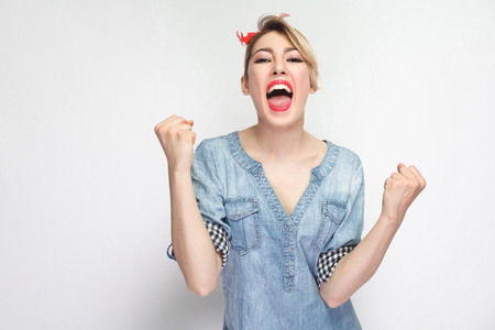 Portrait of happy cheering beautiful young woman in casual blue denim shirt with makeup and red headband standing, screaming and celebraiting her victory. studio shot, isolated on white background.の写真素材