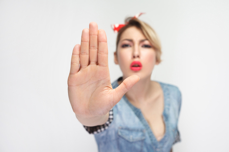 Do not come to me. Portrait of serious beautiful young woman in casual blue denim shirt with red headband standing and looking at camera with stop gesture. studio shot, isolated on white background.の写真素材
