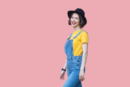 Portrait of happy beautiful young woman in yellow t-shirt and blue denim overalls with makeup and black hat standing and looking at camera with toothy smile. studio shot isolated on pink background.の写真素材