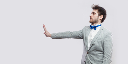 Profile side view portrait of serious handsome bearded man in casual grey suit, blue bow tie standing and looking straight with stop hand gesture sign. studio shot, isolated on light grey background.の写真素材