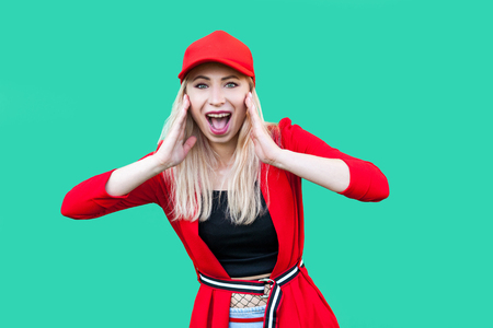Portrait of amazed beautiful blond young hipster woman in red blouse and cap, standing, touching her face and looking at camera with suprised face. indoor studio shot, isolated on green background.の写真素材