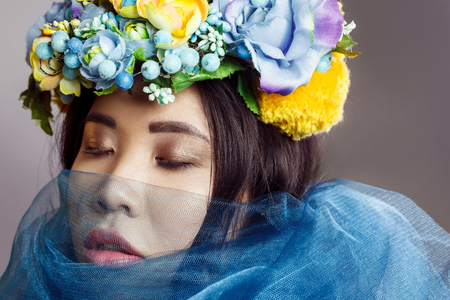 portrait of attractive asian woman with floral hat and blue veil posing with closed eyes on light grey background . indoor studio shot.の写真素材