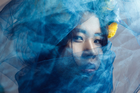 portrait of sensual gorgeous asian woman looking at camera through blue veil on face . indoor studio...