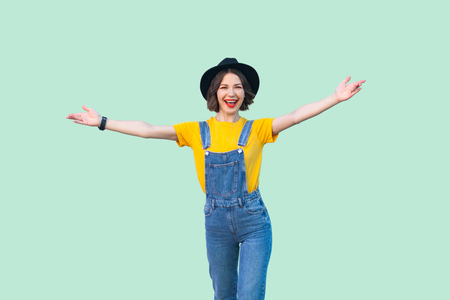 Portrait of happy pretty young girl in blue denim overalls, yellow shirt, black hat standing with raised arms and looking at camera with toothy smile. studio shot isolated on light green background.の写真素材