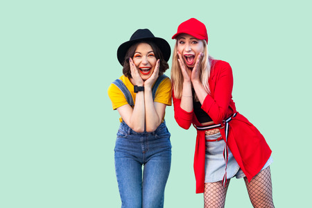 Portrait of two best friend beautiful amazing happy fashionable hipster girls standing and screaming with unbelievable face, looking at camera. Indoor studio shot, isolated on green background.の写真素材