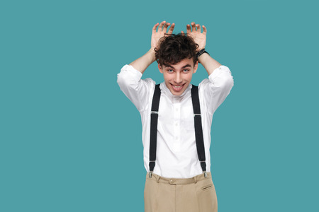 Funny man with bunny ears gesture and tongue out looking at camera. Portrait of handsome hipster curly young businessman in classic casual white shirt standing. studio shot isolated on blue backgroundの写真素材