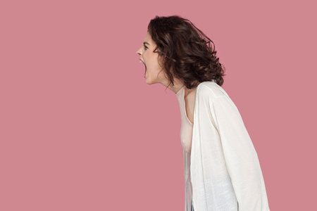 Profile side view portrait of brunette young woman with curly hairstyle in casual style standing and screaming. people expression and emotion. indoor studio shot isolated on pink background.の写真素材