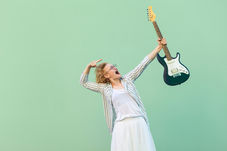 Portrait of young blonde woman in white shirt, skirt, and striped blouse with eyeglasses standing holding and pointing on her electric guitar. indoor studio shot isolated on light green background.の写真素材
