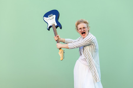 Portrait of angry young blonde woman in white striped blouse with eyeglasses standing, holding electric guitar looking, screaming and attacking. indoor studio shot isolated on light green background.の写真素材