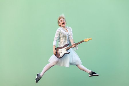 Portrait of young happy blonde woman in white shirt, skirt, and striped blouse with eyeglasses holding electric guitar, screaming and jumping. indoor studio shot isolated on light green background.の写真素材