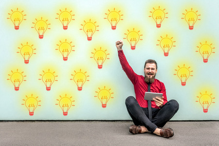 Happy bearded young businessman in red shirt holding tablet sitting on floor, celebrating amazing idea or victory, looking at camera. Yellow bulb lamp drawed on light green background. studio shot.の写真素材