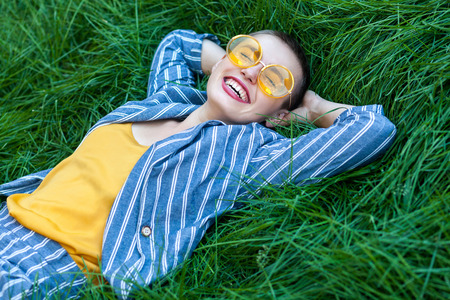 Portrait of Happy fancy young woman with short hair in casual blue striped suit, yellow shirt, glasses lying down on green grass, holding hands behind head, relaxing with toothy smile. summertime shotの写真素材