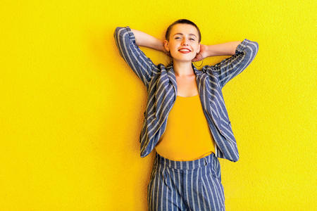 Portrait of happy young short hair beautiful woman in yellow shirt and striped suit standing, holding her head and looking at camera with toothy smile. indoor studio shot isolated on yellow backgroundの写真素材