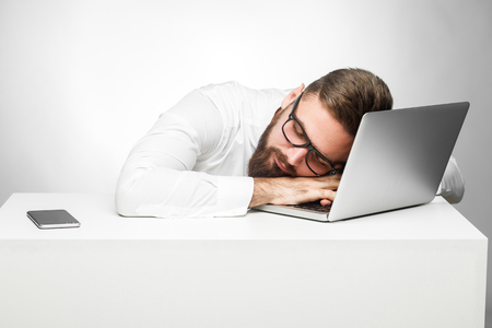 Sweet dreams in the work station. Portrait of sleepy tired freelancer in white shirt are sitting in office is snoozing at his work place near laptop. Indoor, studio shot, gray background, isolatedの写真素材