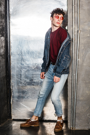 Full length portrait of stylish young fashion model man in bright red sunglasses and denim casual style posing near metallic door. lean and looking away. indoor, studio shot.の写真素材