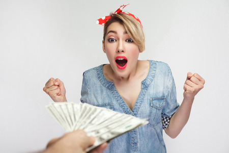 Surprised beautiful young woman in casual denim shirt and red headband standing with raised arms and unbelievable big eyes, won a lot of money. indoor studio shot, isolated on white backgroundの写真素材
