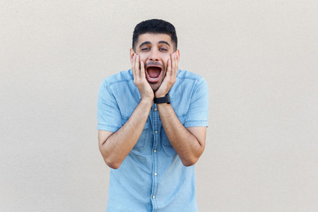 Portrait of surprised handsome young bearded man in blue shirt standing, touching his face and looking at camera with amazed face. indoor studio shot isolated on light beige wall background.の写真素材