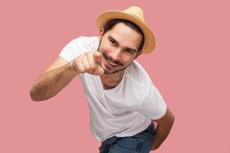 Portrait of funny bearded young man in white shirt with hat standing, pointing and looking at camera...