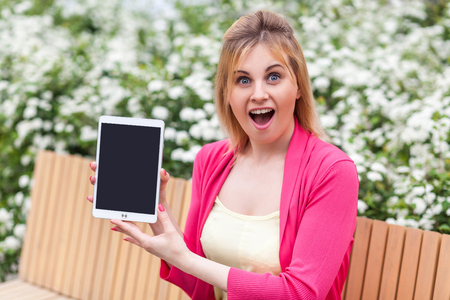 It1s your? Portrait of shocked beautiful young businesswoman in casual style sitting in bench on park, showing tablet empty screen with unbelievable face, opened mouth and looking at camera. Outdoorの写真素材