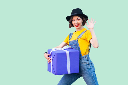 Cheerful happy young girl in hipster wear in denim overalls and black hat standing and trying to...