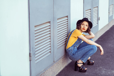 young woman in blue denim overalls and yellow tshirt with black hat sensual looking up while posing...