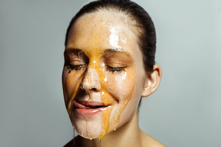 Portrait of beautiful young brunette woman with freckles and honey on face, tongue out and licking with closed eyes and passion. indoor studio shot isolated on gray background.の写真素材