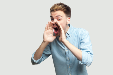 Portrait of handsome young man in light blue shirt standing with hand near mouth and screaming to share something. indoor studio shot on grey background copy space.の写真素材