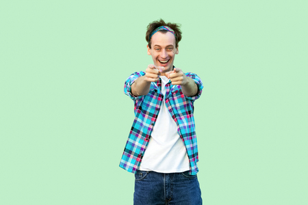 Portrait of excited young man in casual blue checkered shirt and headband standing with toothy smile, looking and pointing at camera. indoor studio shot, isolated on light green background.の写真素材