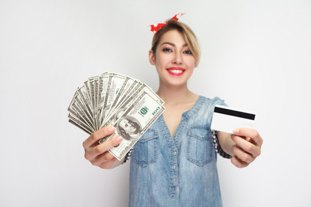 Make choice for usability cash or card. Young girl in casual blue denim shirt standing, demonstrate cash and credit card for choosing, toothy smile. indoor studio shot, isolated on white backgroundの写真素材