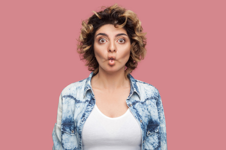Portrait of funny young woman with curly hairstyle in casual blue shirt standing with fish pout lips and looking at camera with big eyes. indoor studio shot, isolated on pink background.の写真素材