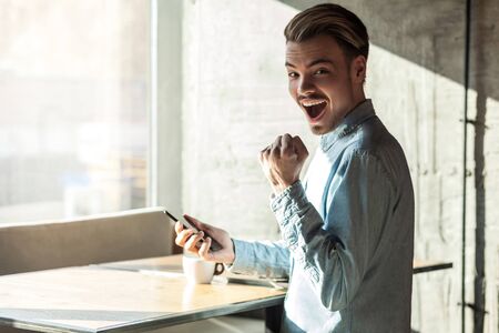Side view portrait of happy success suprised bearded young man in denim blue shirt standing holding phone, looking at camera and celebrate victory with fist. Indoor, lifestyle, window backgroundの写真素材