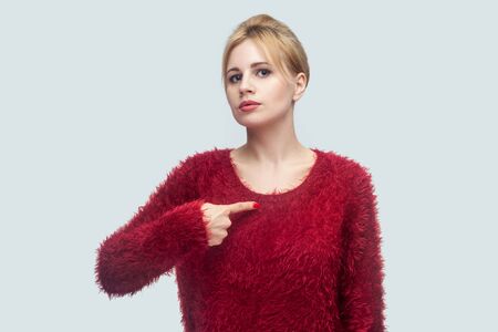 Portrait of serious proud beautiful young blond woman in red blouse standing, pointing herself and looking at camera with confident face. indoor studio shot isolated on light gray background.の写真素材