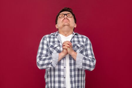 Oh god please help me. Portrait of hopeful sad worry middle aged man in casual checkered shirt and eyeglasses standing, praying and looking at up. indoor studio shot, isolated on dark red background.の写真素材
