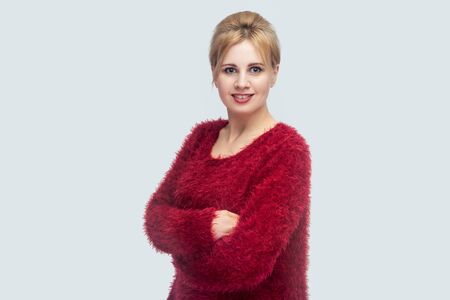 Portrait of happy beautiful young blond woman with makeup and bun hair, red blouse standing, crossed arms and looking at camera with toothy smile. indoor studio shot isolated on light gray background.の写真素材