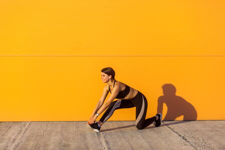 Side view of young beautiful sporty yogi female in black sportwear stretching on the street floor standing on knee, looking straight, Outdoor, orange wall background, sport and healthy conceptの写真素材