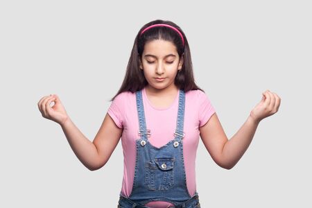 Yoga. Portrait of calm relaxed beautiful brunette young girl in casual style, pink t-shirt and blue denim overalls standing, raised arms and meditating. studio shot, isolated on light gray background.の写真素材