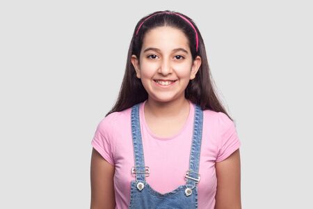 Portrait of happy beautiful brunette young girl in casual style, pink t-shirt and blue denim overalls standing and looking at camera with toothy smile. indoor studio shot, isolated on gray background.の写真素材