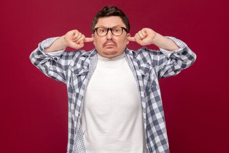 Portrait of serious middle aged business man in casual checkered shirt, eyeglasses standing, putting fingers in ears and dont want to listen anything more. studio shot, isolated on dark red backgroundの写真素材
