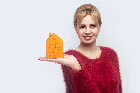 woman in red blouse standing, holding paper small house and propose you to planning new home to buy. Indoor, isolated, studio shot, gray backgroundの写真素材