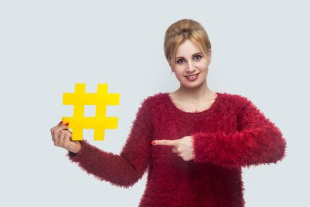 Portrait of happy young woman in red blouse standing and holding large big yellow hashtag sign and pointing finger, social media concept. Indoor, isolated, studio shot, copy space, gray backgroundの写真素材