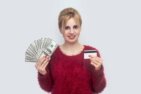 Happy cheerful beautiful young businesswoman in red blouse standing, holding many fan of dollars,...