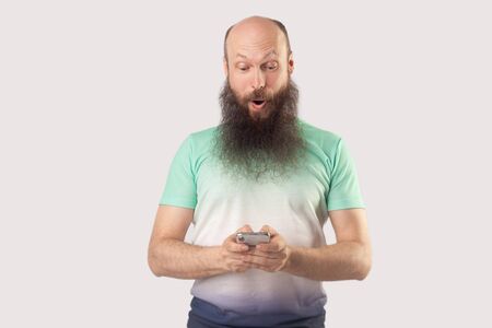 Portrait of shocked middle aged bald bearded man looking at mobile smart phone display with surprised face. reading good news or something amazing. indoor studio shot, isolated on grey background.の写真素材