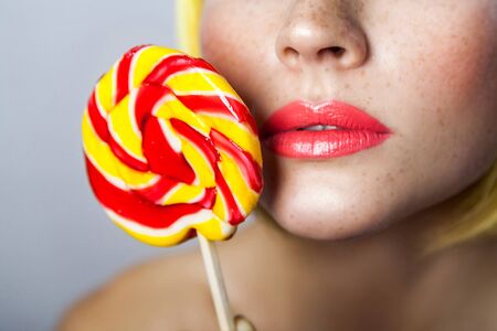 Closeup beauty portrait of cute young female model with freckles, red makeup and yellow wig, holding colorful candy stick near her red beautiful lips. indoor studio shot, isolated on gray background.の写真素材