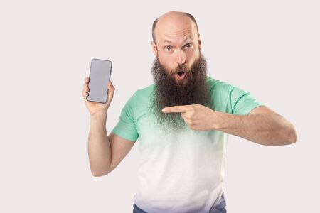 Portrait of shocked middle aged bald bearded man standing with mobile smart phone pointing at empty screen and looking with surprised face. indoor studio shot, isolated on grey background.の写真素材