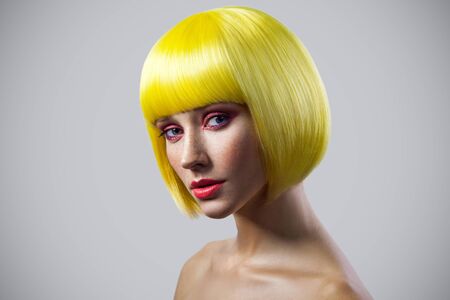 Beauty portrait of calm cute young female model with freckles, red makeup and yellow wig looking at camera with serious face. indoor studio shot, isolated on gray background.の写真素材